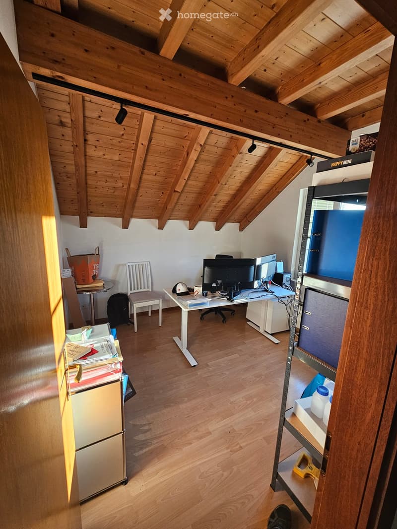A furnished room with wooden beams on the ceiling, a desk with a laptop and screen, a chair, a small table with a cup, and a shelf with various items.