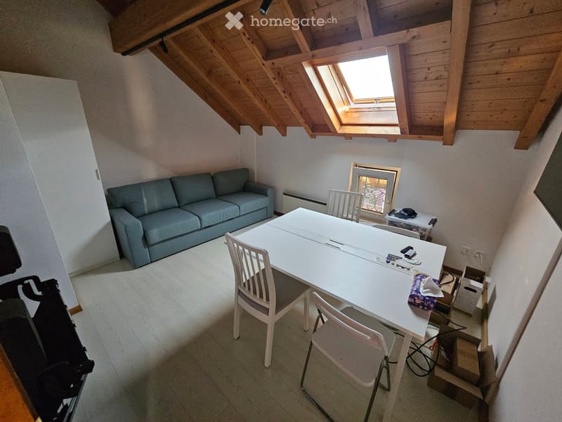 A furnished attic room with a grey sofa, a white dining table, and chairs, featuring exposed wooden beams and a skylight window.