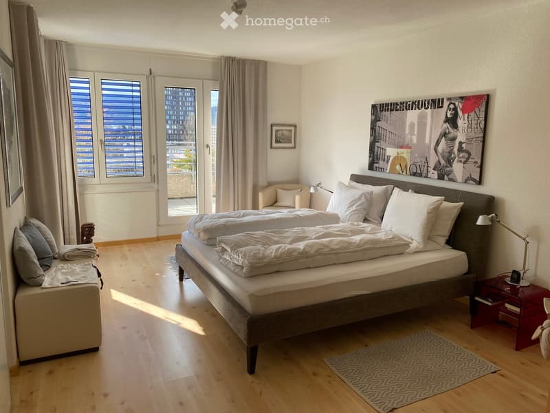 Modern bedroom with a large double bed, nightstand, chair, and artwork on the wall. The room features wooden flooring, large windows with blinds, and a cozy seating area.