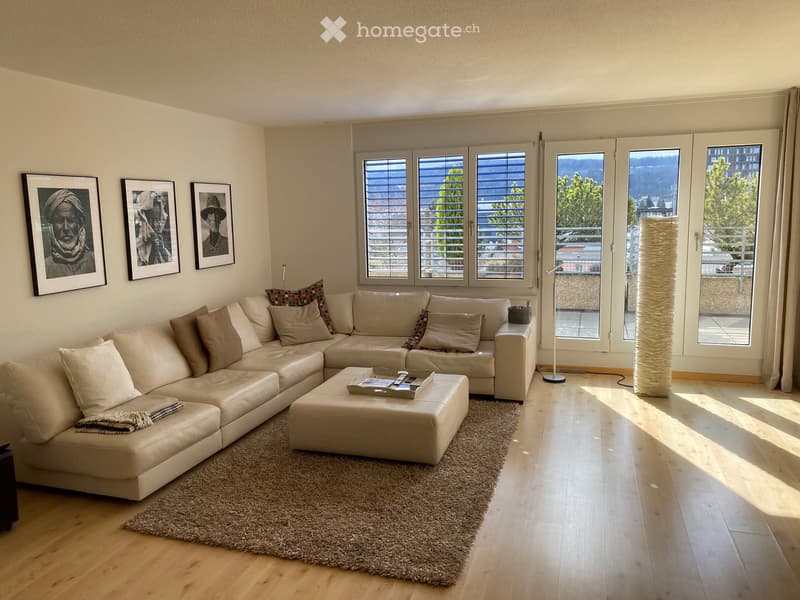 Modern living room with a large sectional sofa, coffee table, ottoman, floor lamp, and artwork on the wall. Large windows offer a scenic view.
