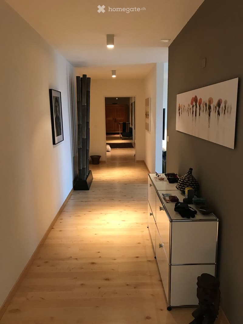 A long, well-lit hallway with wooden flooring, white walls, and modern decor including a glass console table, artwork, and a sculpture on the left side.