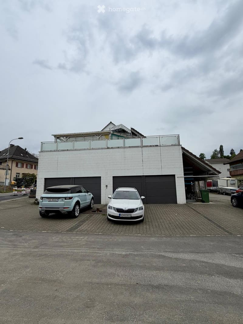Modern two-story house with white exterior, large garage, and balcony on the upper level. Two cars are parked in front of the garage.