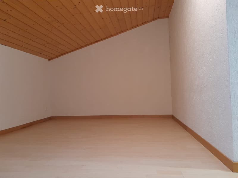 Empty room with light wooden flooring and white walls, featuring a slanted wooden ceiling.