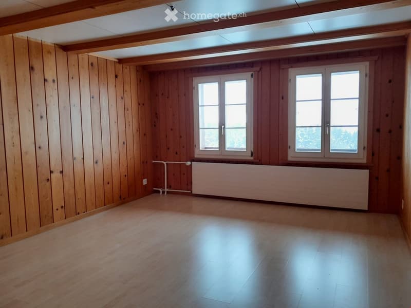 Empty room with wooden walls and floor, large windows, radiator, and exposed wooden beams on the ceiling.