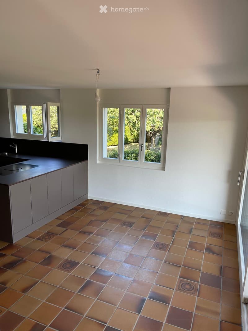 Empty kitchen with tiled floor, grey cabinets, and a black countertop. Windows offer a view of greenery outside.