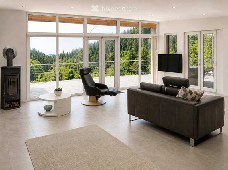 Modern living room with large windows offering a scenic view, featuring a dark leather sofa, a white coffee table, a black leather chair, a fireplace, and a television.