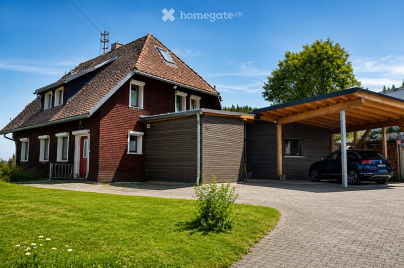 Two-story house with red brick exterior, white-framed windows, and a covered driveway leading to a garage. A modern carport shelters a parked vehicle.