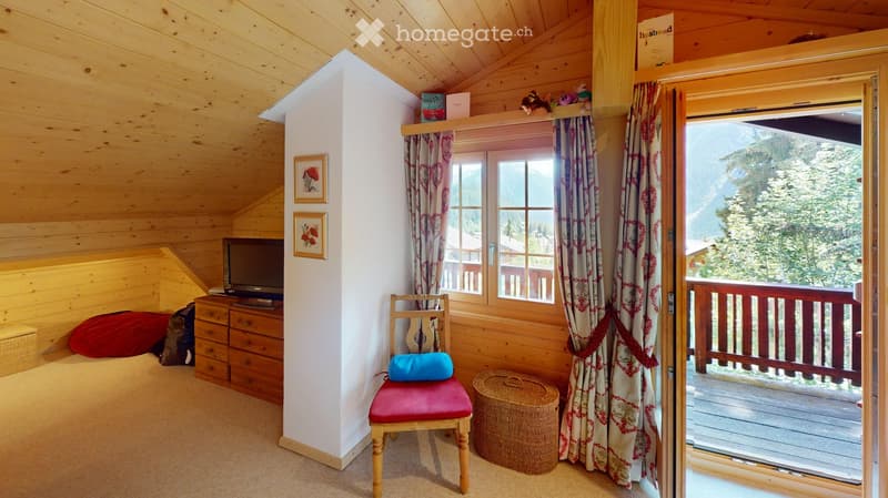 Cozy wooden bedroom with slanted ceiling, large window with view, red cushioned chair, TV, and decorative items.