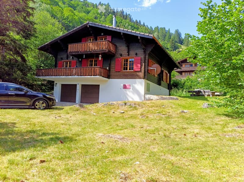 A two-story wooden house with red shutters, a balcony, and a garage, situated in a grassy area surrounded by trees.