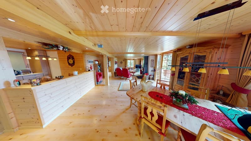 A spacious, well-lit living and dining area with wooden ceilings and floors, featuring a dining table with chairs, a sofa, a television, and a kitchen counter.