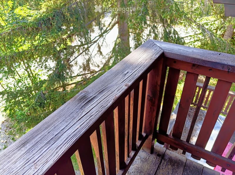 Wooden balcony railing with vertical slats and a smooth top rail, surrounded by lush green trees.