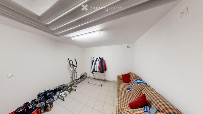A small, furnished room with a plaid-patterned sofa, exercise bike, clothing rack, and several bags on the floor.