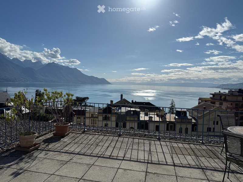 Balcony with a view of a lake and mountains, featuring a tiled floor, metal railing, potted plants, and outdoor furniture.