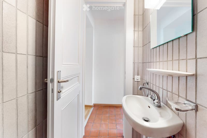 A small bathroom with a white sink, chrome faucet, a mirror with a green frame, and a white shelf above the sink. The walls are tiled in beige, and the floor is covered with orange tiles.