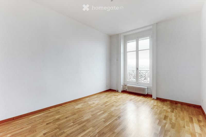 Empty room with white walls, wooden floor, and large window with decorative metal grille. Radiator visible under window.
