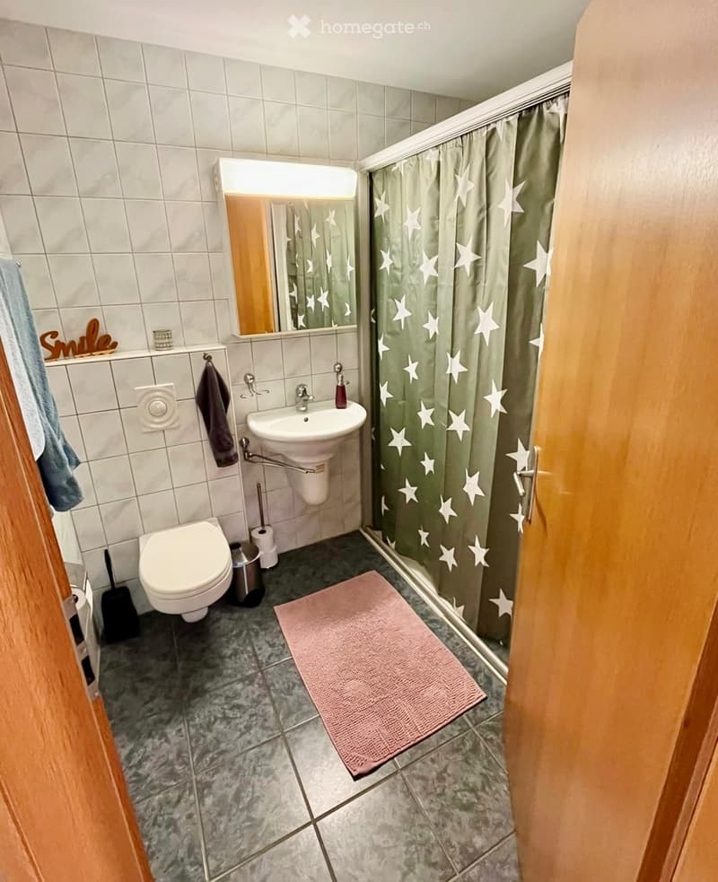 A small bathroom with a toilet, sink, mirror, and shower area with a green star-patterned curtain. Includes a pink bath mat, towels, and a toilet paper holder.