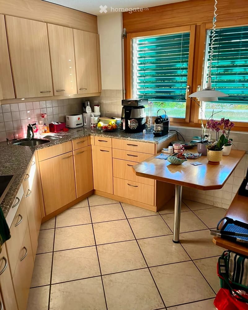 Bright kitchen with light wooden cabinets, granite countertops, and tiled floor. Features a coffee maker, toaster, sink, and a small table with a flower pot.