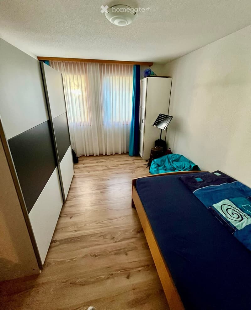 A small bedroom with wooden flooring, a single bed with blue bedding, a wardrobe, and a window with white curtains.
