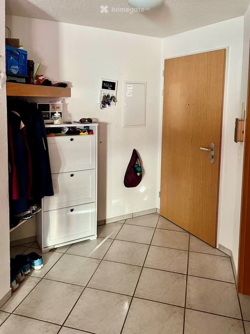 Hallway with tiled floor, white walls, coat rack, shoe rack, and open wooden door leading to another room.