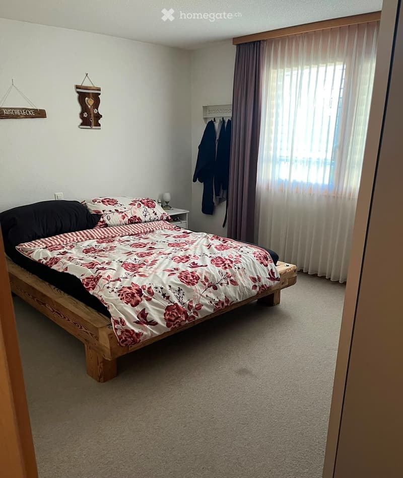 Cozy bedroom with a wooden bed frame, floral duvet, nightstand, and large window with curtains.