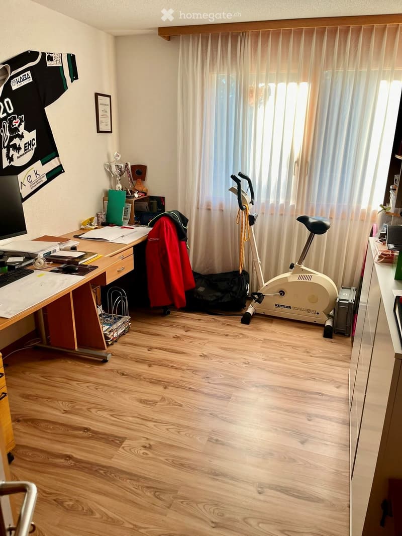 A well-lit room with wooden flooring, a desk with a computer setup, an exercise bike, and various personal items including a framed picture and a trophy on a shelf.
