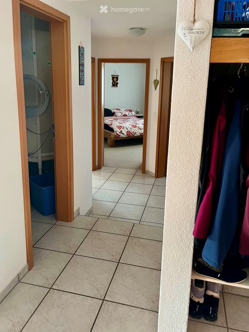 Hallway with tiled floor, wooden doors, and a mirror reflecting a bedroom with a bed and colorful bedding. A coat rack with jackets and shoes is visible on the right side.