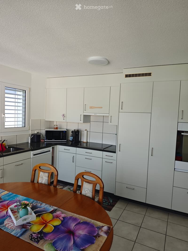 Modern kitchen with white cabinets, stainless steel appliances, and a wooden dining table with chairs. The table is set with a floral table runner and a small vase of flowers.