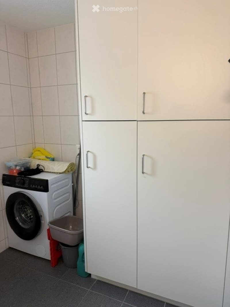 A small bathroom with a washing machine, white cabinets, and a small trash bin.