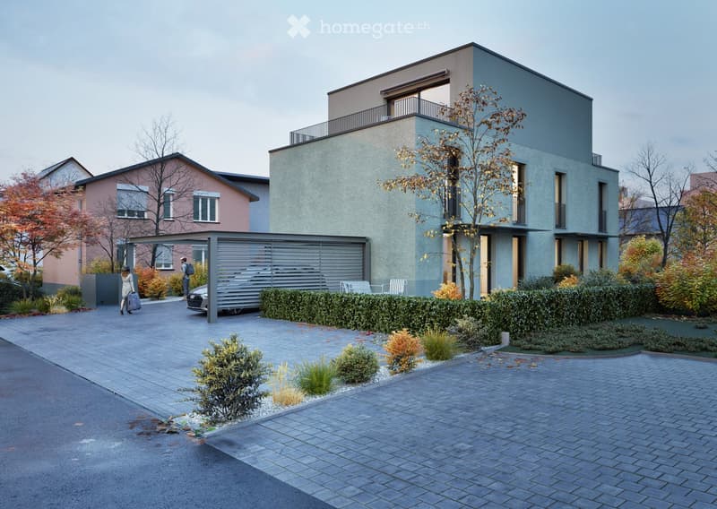 Modern two-story house with grey facade, large windows, and a balcony on the upper floor. The house is surrounded by a well-maintained garden with shrubs and trees, and a paved driveway leads to a covered parking area.