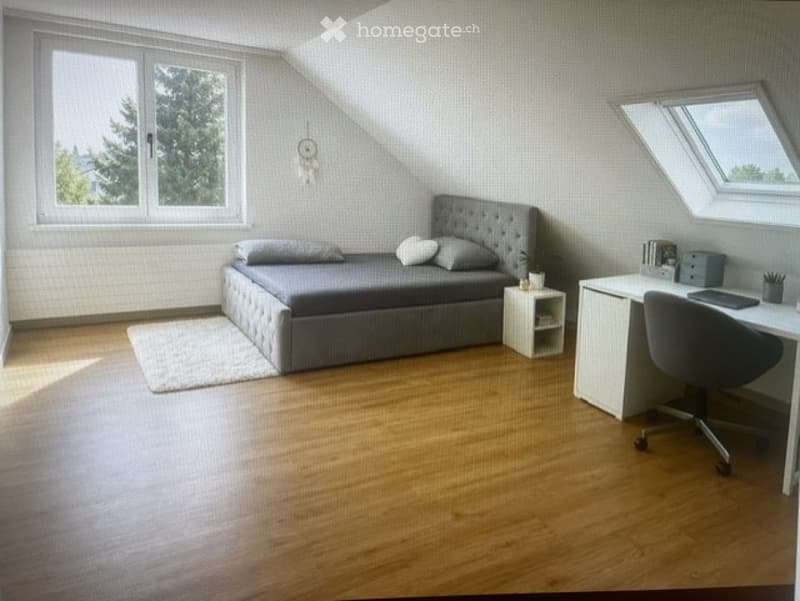 Cozy attic bedroom with wooden flooring, grey upholstered bed, nightstand, desk with chair, laptop, and small rug.