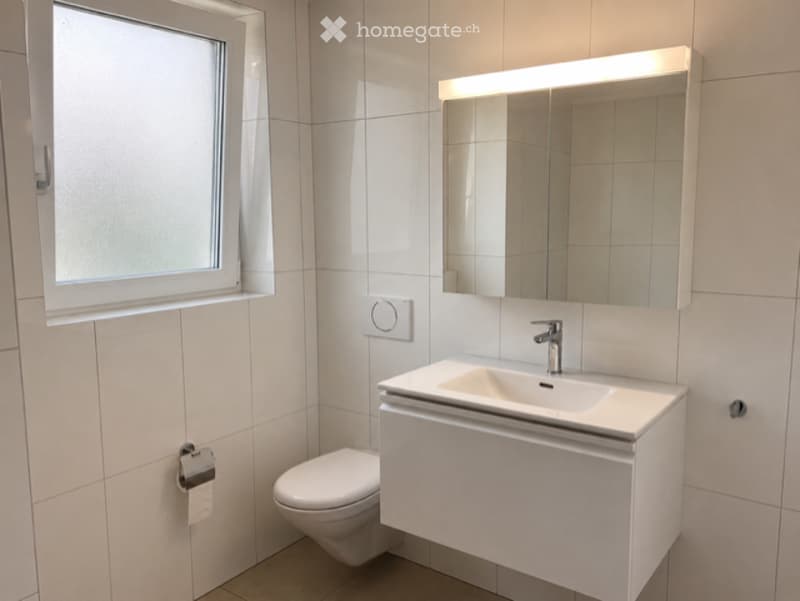 Modern bathroom with white tiles, a large mirror with built-in lighting, a sink with a sleek faucet, and a toilet. The window provides natural light.