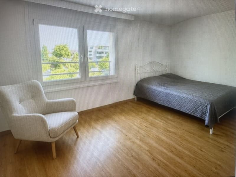 Bright bedroom with wooden flooring, large windows, a single bed with dark bedding, and a light-colored armchair.