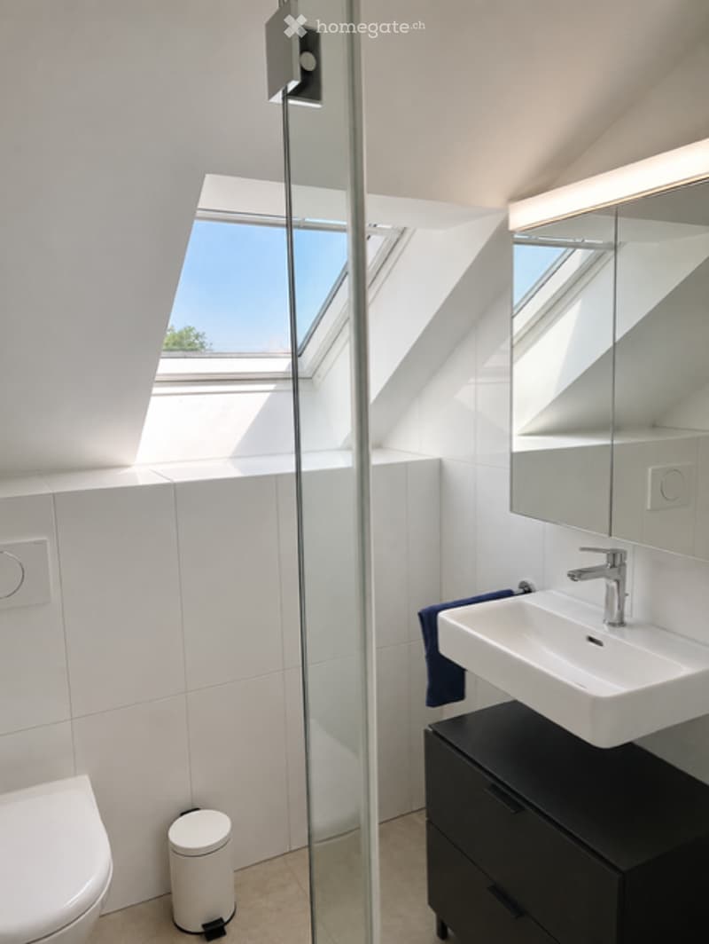 Modern bathroom with white tiles, a large window, a glass shower enclosure, a white sink with a mirror, a black cabinet, and a toilet.