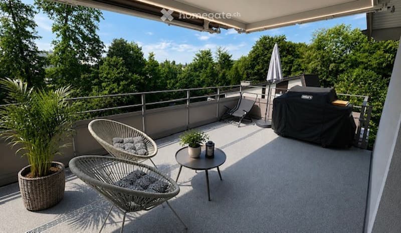 A modern, covered balcony with a view of greenery, featuring wicker chairs, a small round table, potted plants, and a covered grill.