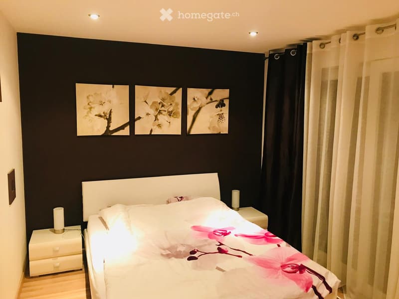 Bedroom with a bed, nightstands, and a wall with three framed pictures. The bed has a white headboard and a floral bedspread. The nightstands have lamps on them. The wall is painted black and has three framed pictures of flowers. The room has a wooden flo