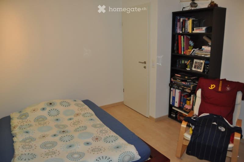Single bed, bedspread, wooden floor, white door, bookshelf, chair with clothes