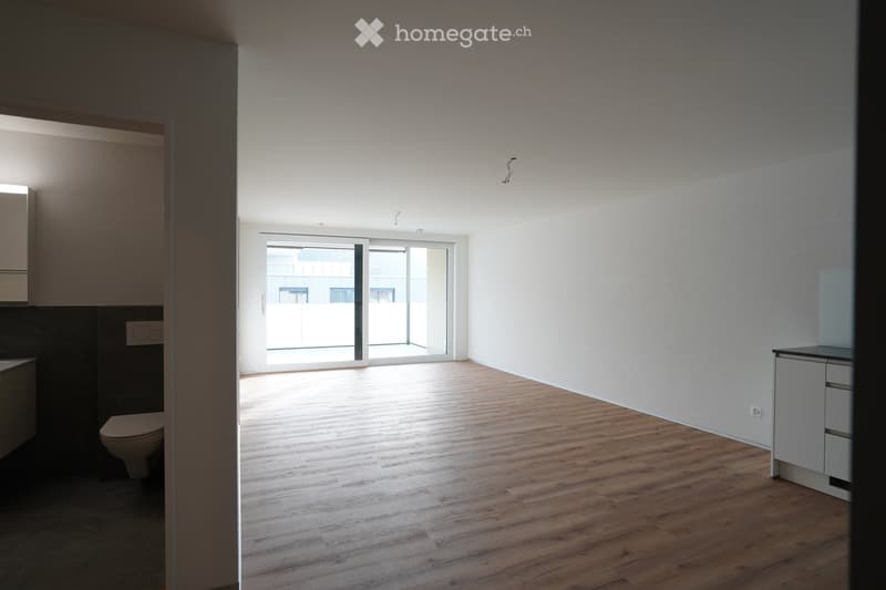 Empty room with wooden flooring, white walls, and a large sliding glass door leading to a balcony. The room includes a small cabinet and a toilet visible in the background.