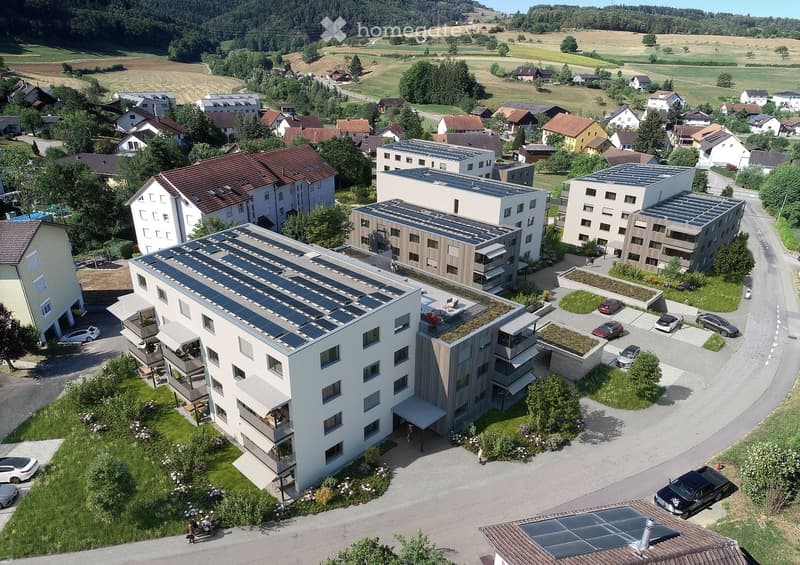 Aerial view of a modern residential complex with multiple buildings, solar panels on rooftops, green spaces, and parking areas.