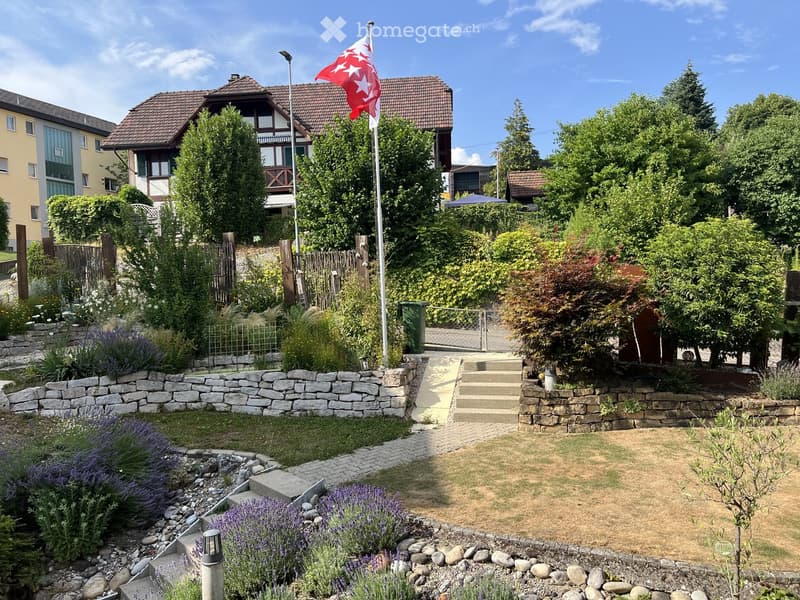 Well-maintained garden with stone walls, lavender bushes, steps leading to a house, and a flagpole with a red and white flag.