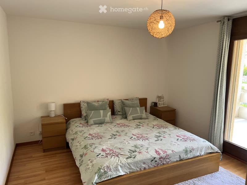 Cozy bedroom with wooden flooring, a neatly made bed with floral bedding, two nightstands with lamps, and a large window with curtains leading to a balcony.