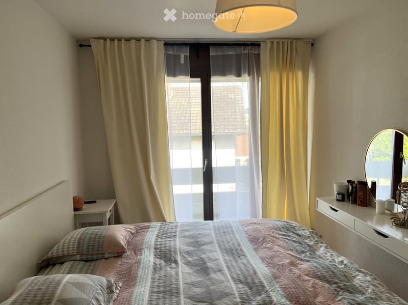 A cozy bedroom with a bed, nightstand, mirror, and a large window with curtains. The room features a modern aesthetic with a patterned bedspread and a sleek white dresser.