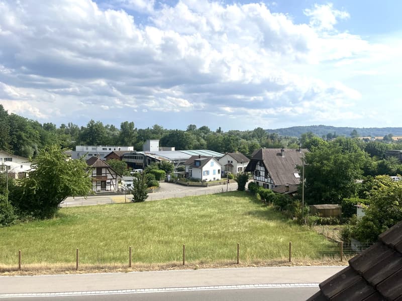 Residential area with houses, trees, and a grassy field under a partly cloudy sky.