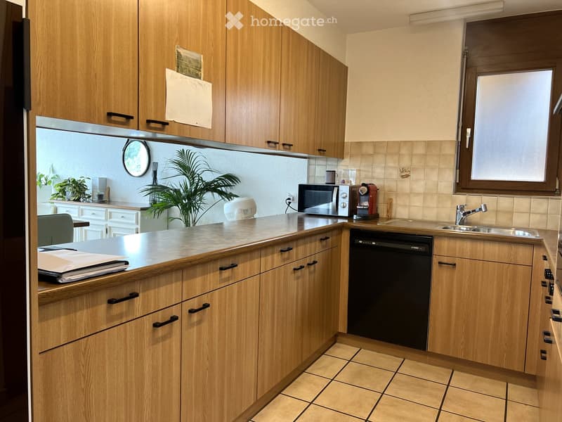 Modern kitchen with wooden cabinets, tiled floor, and stainless steel appliances including a sink, microwave, and coffee maker. A small plant and a mirror are visible.