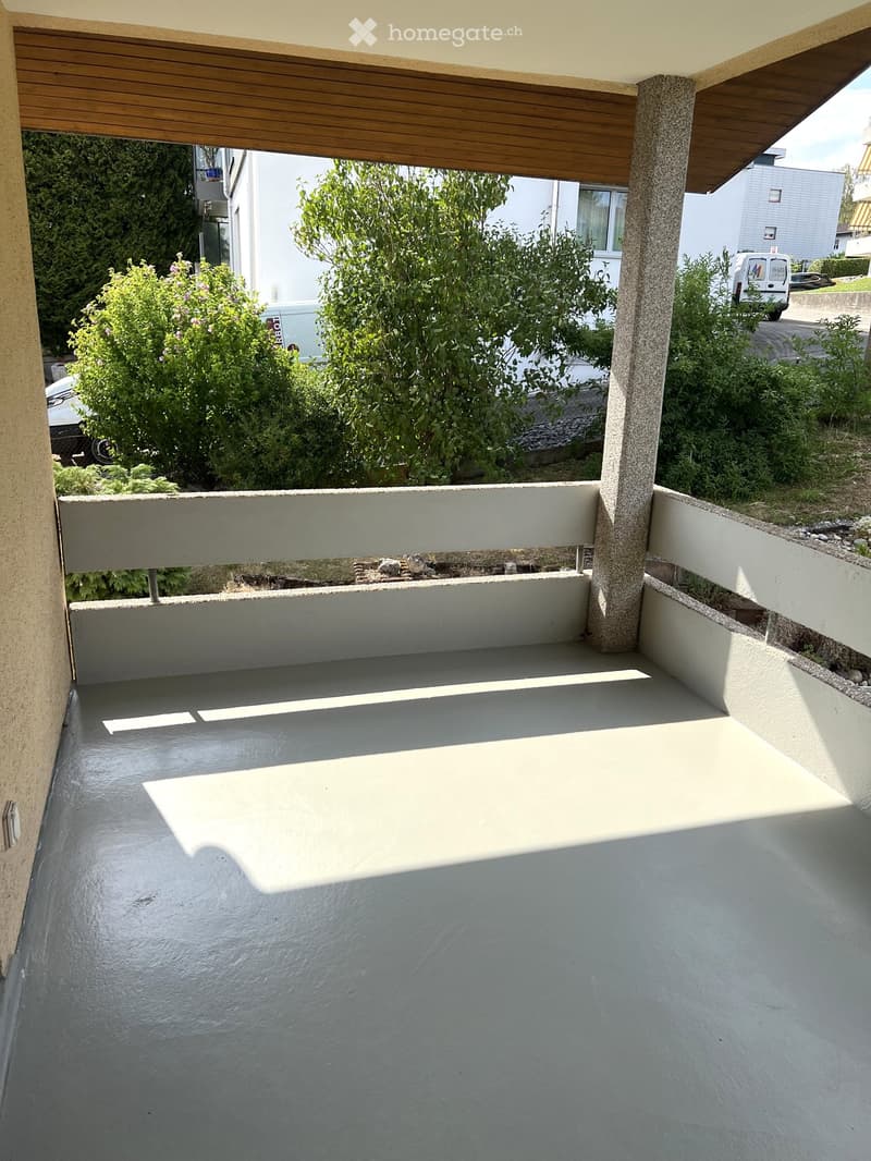 Empty balcony with grey concrete floor, white railing, and wooden ceiling supported by a concrete pillar. The view shows greenery and neighboring buildings.