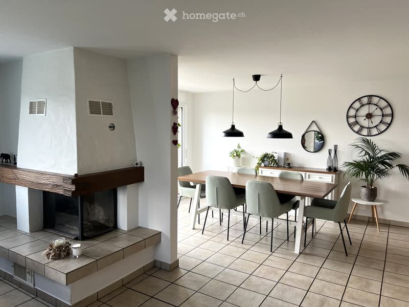 Modern living and dining area with a fireplace, dining table, chairs, and decorative items including a large wall clock and a potted plant.