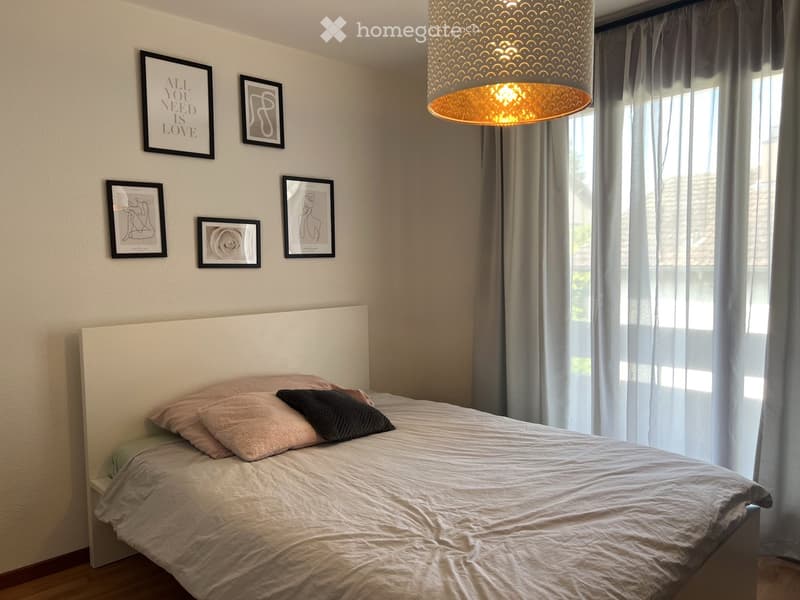 A modern bedroom with a large bed, headboard, pillows, and a hanging lamp. The walls are decorated with several picture frames, and sheer curtains cover the windows.