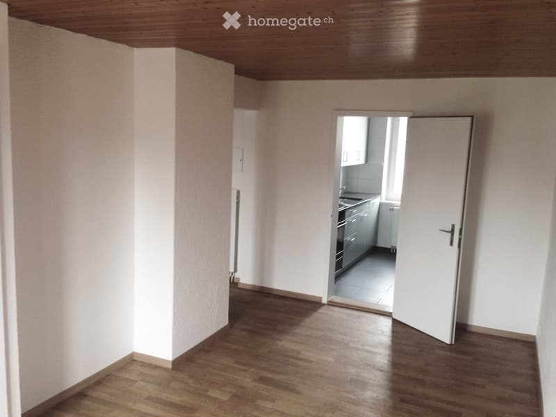 Empty room with wooden flooring and white walls, featuring an open doorway leading to a small kitchen area with cabinets and appliances.
