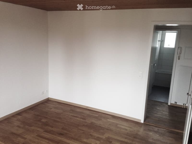 Empty room with wooden flooring and white walls, featuring a doorway leading to a small bathroom with a bathtub and tiled walls.