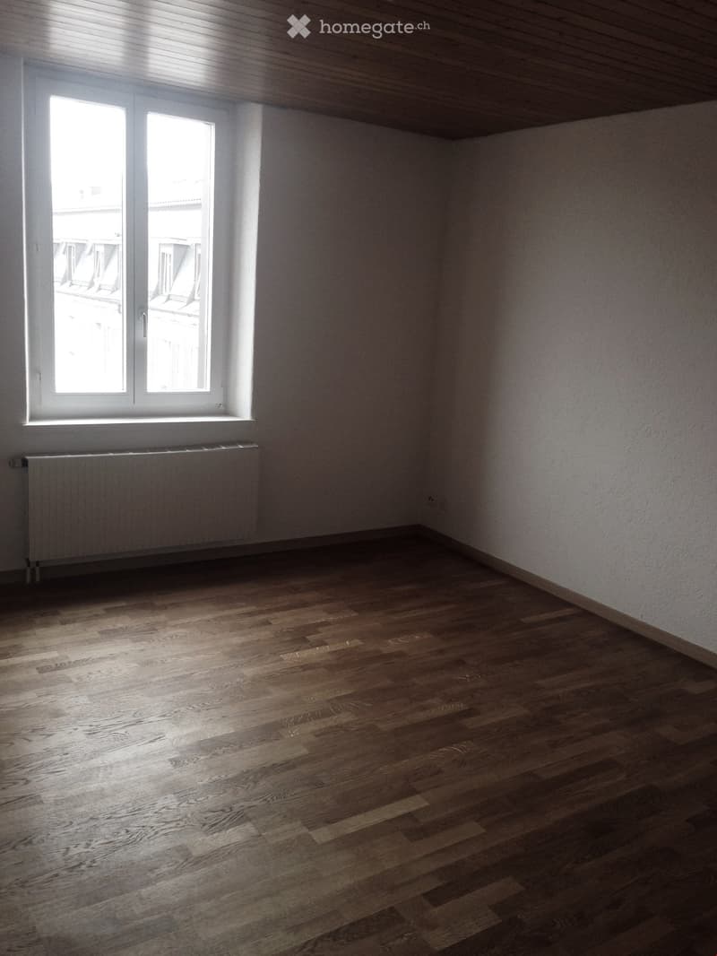 Empty room with wooden flooring, white walls, and a large window with a radiator below it.
