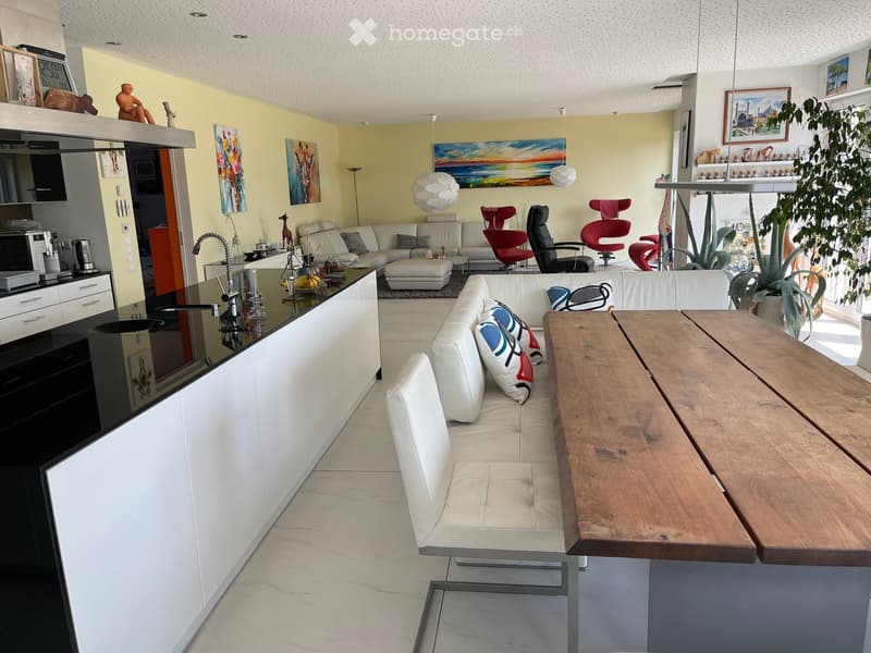 Open-plan living and dining area with white leather sofa, wooden dining table, modern kitchen, and colorful artwork.
